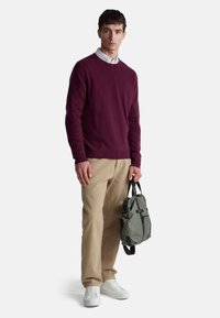 Bordowy sweter z dzianiny, beżowe spodnie i białe trampki; niesie zieloną torbę z czarnymi akcentami i uchwytami. Model stoi na tle białego tła.