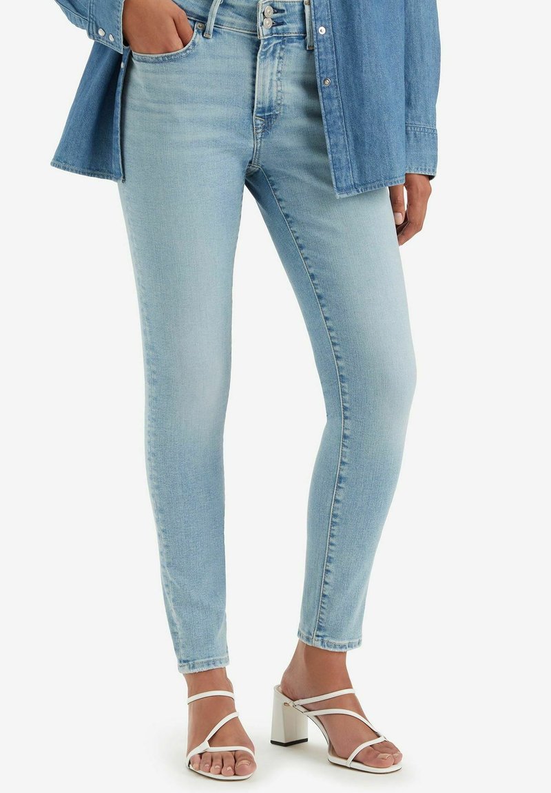 Ljusblå skinny jeans i denimmaterial, med hög midja och subtila blekningar. Kombineras med vita, remförsedda sandaler med blockklack.