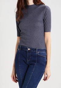 Harmaa ribbineulottu toppi, jossa on lyhyet hihat ja korkea pääntie, yhdistetty tummansinisiin istuviin farkkuihin, joissa näkyy etunappien yksityiskohdat.