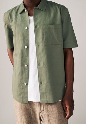 Homme portant une chemise à manches courtes olive verte ouverte sur un t-shirt blanc et un pantalon beige, sur fond uni.