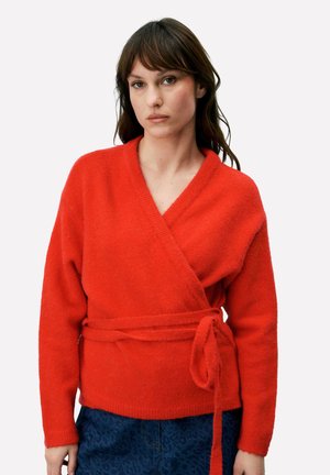 Femme aux cheveux bruns portant un pull cache-cœur rouge vif noué à la taille et un pantalon bleu foncé à motifs, debout devant un fond uni.