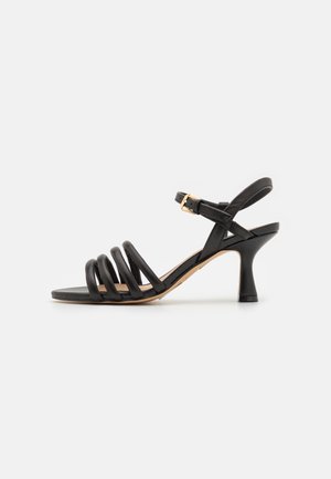 Sandale noire à talon avec quatre brides avant rembourrées, bride de cheville avec boucle dorée, et talon évasé de hauteur moyenne sur fond blanc.