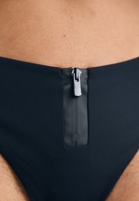 Juodas maudymosi šortai su priekiniais užtrauktukais ir lygia, matinė danga. Dizainas yra minimalistinis ir stilingas, glaudžiai priglundantis prie kūno.