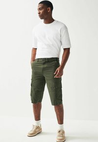 Pantaloni scurți cargo verzi cu buzunare laterale, asortați cu un tricou alb cu guler rotund și încălțări bezhă, purtați cu o curea asortată.