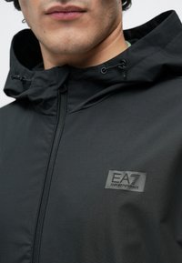 Fekete atlétikai dzseki cipzáras záródással, állítható kapucnival és EA7 logóval ellátott felvarróval. Simaszövetű és tartós anyagból készült, aktív használatra tervezve.
