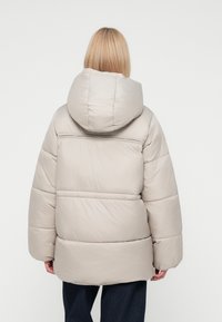 Chaqueta acolchada beige con una gran capucha redondeada, diseño acolchado, grueso acolchado y un corte recto. Presenta un detalle de costura en la parte posterior.