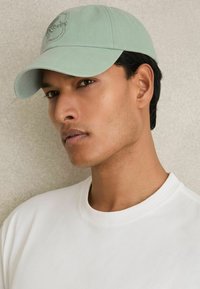Mint green cap with embroidered logo, featuring a curved brim. Worn with a crew-neck white T-shirt against a neutral background.