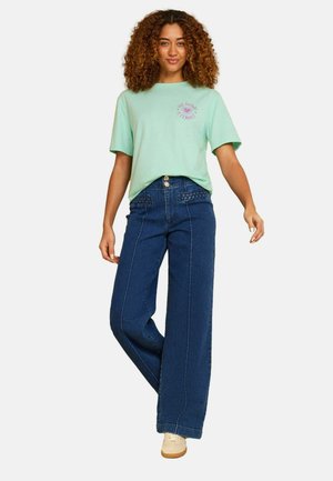 Woman with curly hair wearing a mint green T-shirt with pink text and dark blue wide-leg jeans with beige sneakers, standing on one leg.