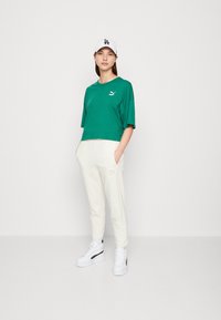 T-shirt verde corto con logo Puma, pantaloni bianchi e sneakers bianchi con accentuazioni nere. Outfit atletico casual su sfondo neutro.