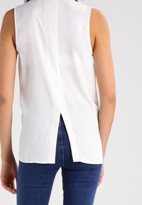 Blusa blanca sin mangas con una abertura en la parte posterior y una costura vertical contrastante que recorre el centro. Tejido suave con una textura lisa.