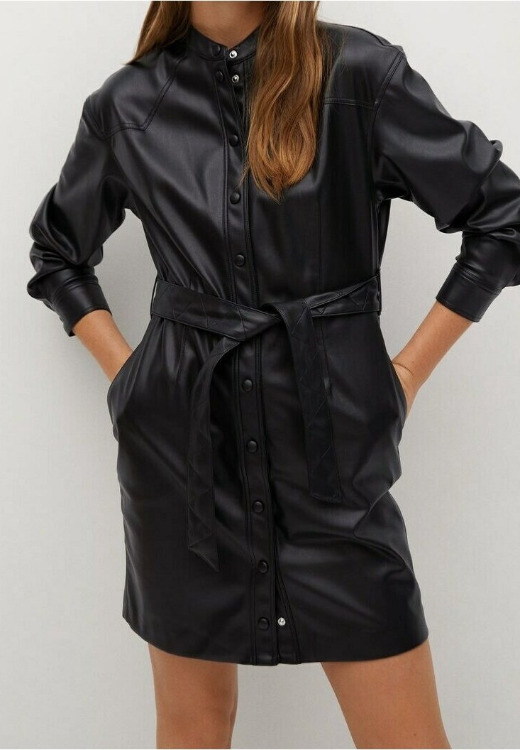 Femme portant une robe boutonnée en similicuir noir à manches longues, ceinturée à la taille avec une ceinture nouée, les mains dans les poches.