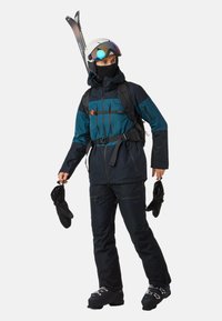 Skikleding met een marineblauwe en teal jas, zwarte broek. Beschikt over een capuchon, ritsen, een rugzak en skibenodigdheden. Handschoenen in de linkerhand gehouden.