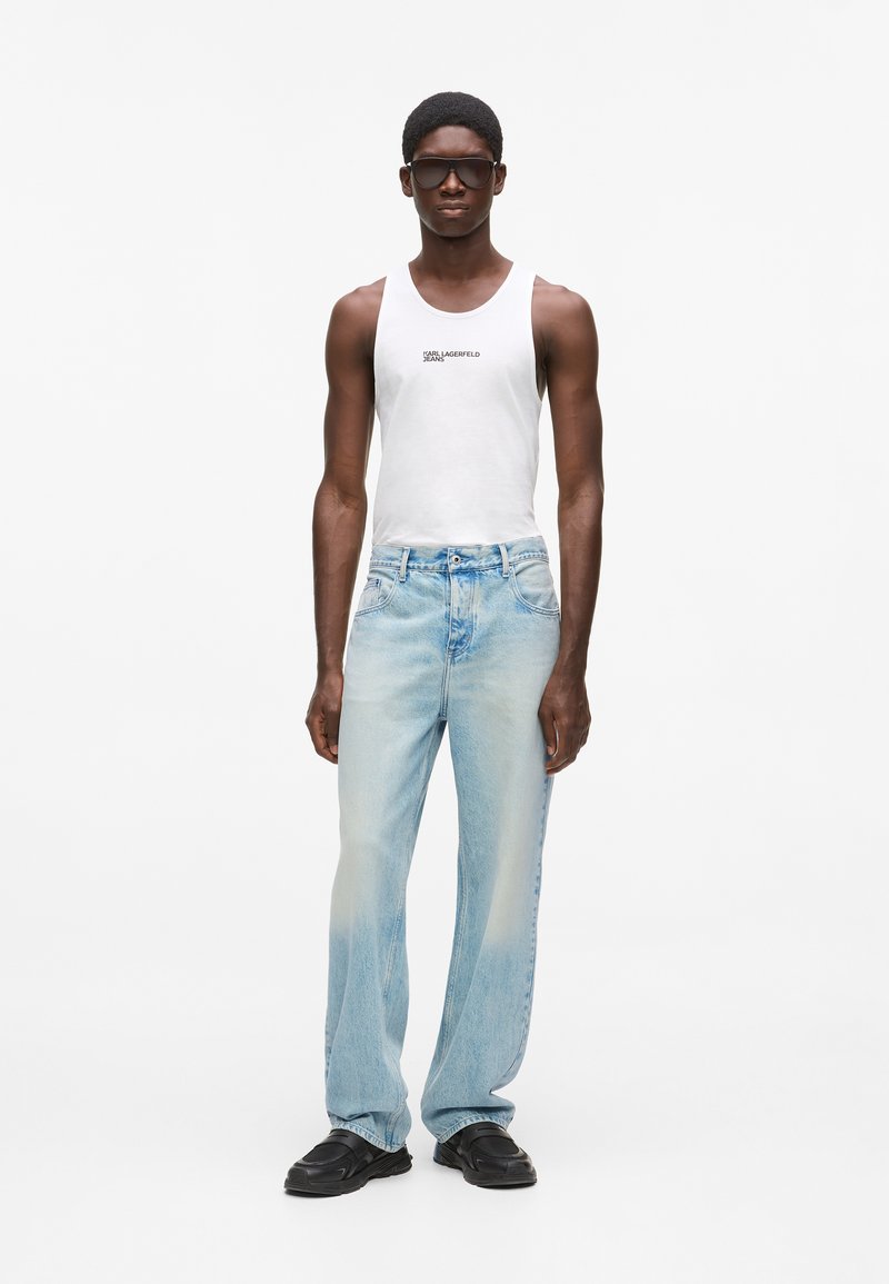 Άνδρας που φοράει μαύρα γυαλιά ηλίου, άσπρο αμάνικο τοπ με το λογότυπο "Karl Lagerfeld Jeans", ανοιχτό μπλε ξεθωριασμένο τζιν και μαύρα μοκασίνια, στέκεται σε λευκό φόντο.