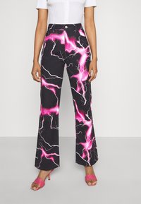 Kvinna iklädd högmidjade svarta jeans med rosa blixtmönster, vit kortärmad topp och rosa klacksandaler.