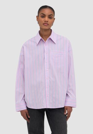 Kobieta w oversize jasnoróżowej koszuli na guziki z cienkimi niebieskimi i białymi paskami oraz czarnych spodniach, stojąca na jednolitym tle.