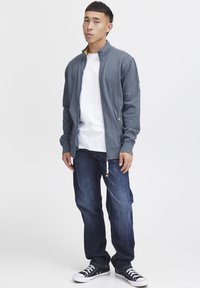 Grå zip-jakke med ribbet krage, hvit t-skjorte, mørkeblå denimjeans og svarte sneakers med hvite såler. Uformelt antrekk, minimal detaljering.