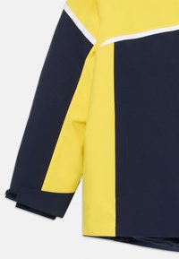 Chaqueta resistente al agua con un diseño en bloques de color amarillo y marino, que presenta rayas acentuadas en blanco y puños de manga ajustables.
