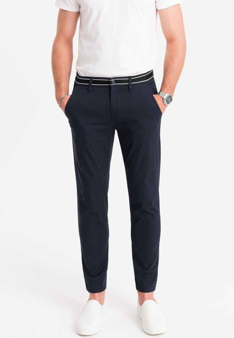 Chino navy realizzati in morbido cotone con vestibilità slim, dotati di una cintura a righe bianche e nere e tasche laterali. Indossati con scarpe bianche.