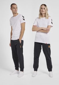 Hvide t-shirts med sorte chevron-mønstre på ærmerne, parret med mørke sweatpants med et orange logo, begge med hvide sneakers.