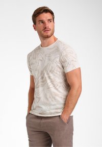 T-shirt léger beige à manches courtes avec un motif abstrait texturé d'une teinte légèrement plus foncée. Encolure ronde avec une silhouette ajustée.