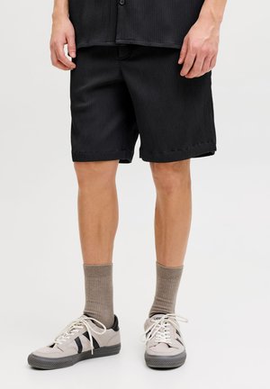 Muž nosi čierne rebrované šortky, béžové ponožky a béžové tenisky s čiernymi pruhmi, stojí na bielom pozadí.