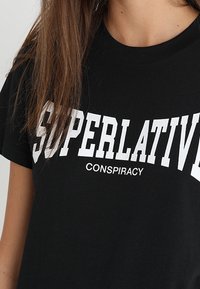 Černé bavlněné tričko s výrazným bílým textem: "SUPERLATIV" v kapitálkách a "KONSPIRACE" v menším písmu pod tím. Krátké rukávy.