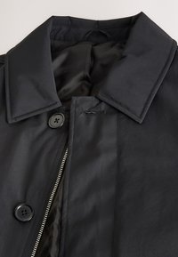 Manteau noir avec large col, deux gros boutons et fermeture éclair argentée visible partiellement ouverte sur un fond clair.