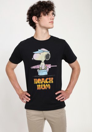 Jonge man draagt een zwart T-shirt met Snoopy die een roze paraplu en blauwe koelbox vasthoudt, tekst "BEACH BUM", staat met handen in de zij.