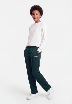 Smilende ung person, der står med hænderne i lommerne, iført hvid langærmet skjorte, mørkegrønne bukser og hvide sneakers på hvid baggrund.