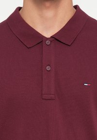 Burgunder farge polo skjorte med klassisk krage, to-knapp knappelukking, og liten tri-farget logo detalj på brystet. Teksturert stoff.