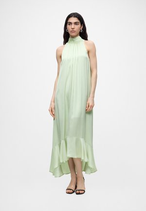 Γυναίκα με αμάνικο midi φόρεμα από σατέν ανοιχτού πράσινου χρώματος με ψηλό λαιμό και βολάν στο τελείωμα, και μαύρα ψηλοτάκουνα ανοιχτά σανδάλια, στεκόμενη μπροστά σε λευκό φόντο.