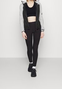 Leggings noirs avec des accents côtelés, associés à un haut court noir et un coupe-vent léger gris clair à capuche, complétés par des baskets foncées.