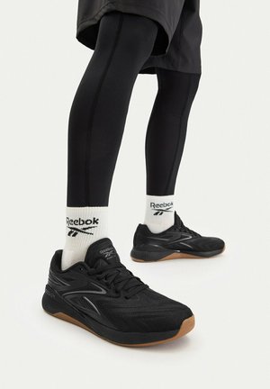 Nohy v černých legínách, bílých ponožkách Reebok a černých běžeckých botách Reebok s gumovou podešví na světlém pozadí.