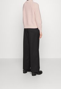 Personne portant un pull en maille rose clair, un pantalon large noir et des bottes noires épaisses, debout de profil sur un fond blanc uni.