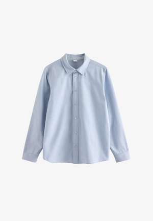 Teenage-Junge lächelt, trägt hellblaues Hemd mit Knöpfen, weißes T-Shirt, lockere blaue Jeans und dunkle Sneakers, geht vor einem schlichten Hintergrund.