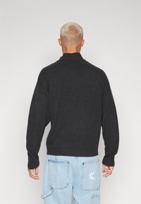 Maglione grigio scuro lavorato a maglia con collo alto, spalle scese e polsini a coste. Indossato con pantaloni cargo azzurri chiaro con tasca applicata.