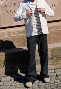 Mężczyzna dostosowujący biały żakiet do pasiastej koszuli, noszący luźne czarne spodnie i beżowe buty, stojący na kamiennej nawierzchni obok engraved wall.