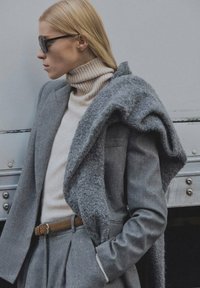 Siv volnen blazer na bež turtlenecku, dopolnjen s sivim teksturiranim šalom, ki je prekrita preko enega ramena. Rjav pas poudarja pas.