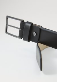 Cinturón de cuero negro con una hebilla metálica de doble punta, que presenta un logo de CK y una textura suave, terminado con un lazo de cuero.