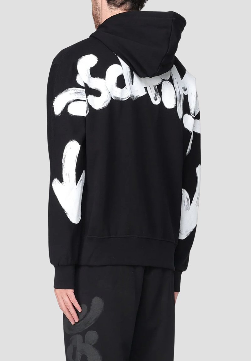 Uomo che indossa un hoodie nero con testo in stile graffiti bianco e frecce sulla schiena e sulle maniche, in piedi contro uno sfondo chiaro e semplice.