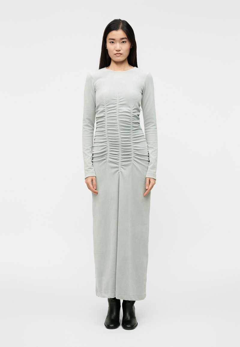 Obleka z dolgimi rokavi, oprijeta, iz svetlo zelene tkanine z naborki spredaj. Obleka sega do členkov, kombinirana s črnimi gležnjarji.