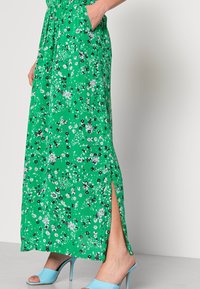 Nainen, joka käyttää vihreää kukkakuosisista maxi-mekkoa, jossa on sivuhalkio, ja sinisiä korkokenkiä avonaisella varrella, käsi taskussa, yksinkertaista taustaa vasten.