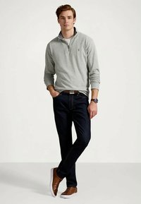 Νεαρός άνδρας στέκεται με το ένα χέρι στην τσέπη, φορώντας ελαφρύ γκρι σκούτερ με φερμουάρ μέχρι το τέταρτο, σκούρα τζιν, καφέ αθλητικά παπούτσια και ρολόι χειρός.