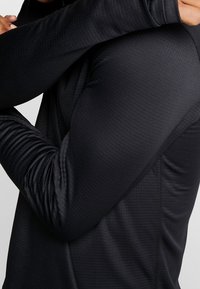 Musta pitkähihainen paita, jossa on teksturoitu kangas, vartalonmyötäinen leikkaus ja tasosaumat. Pyöreä kaula-aukko ja istuvat hihansuut.