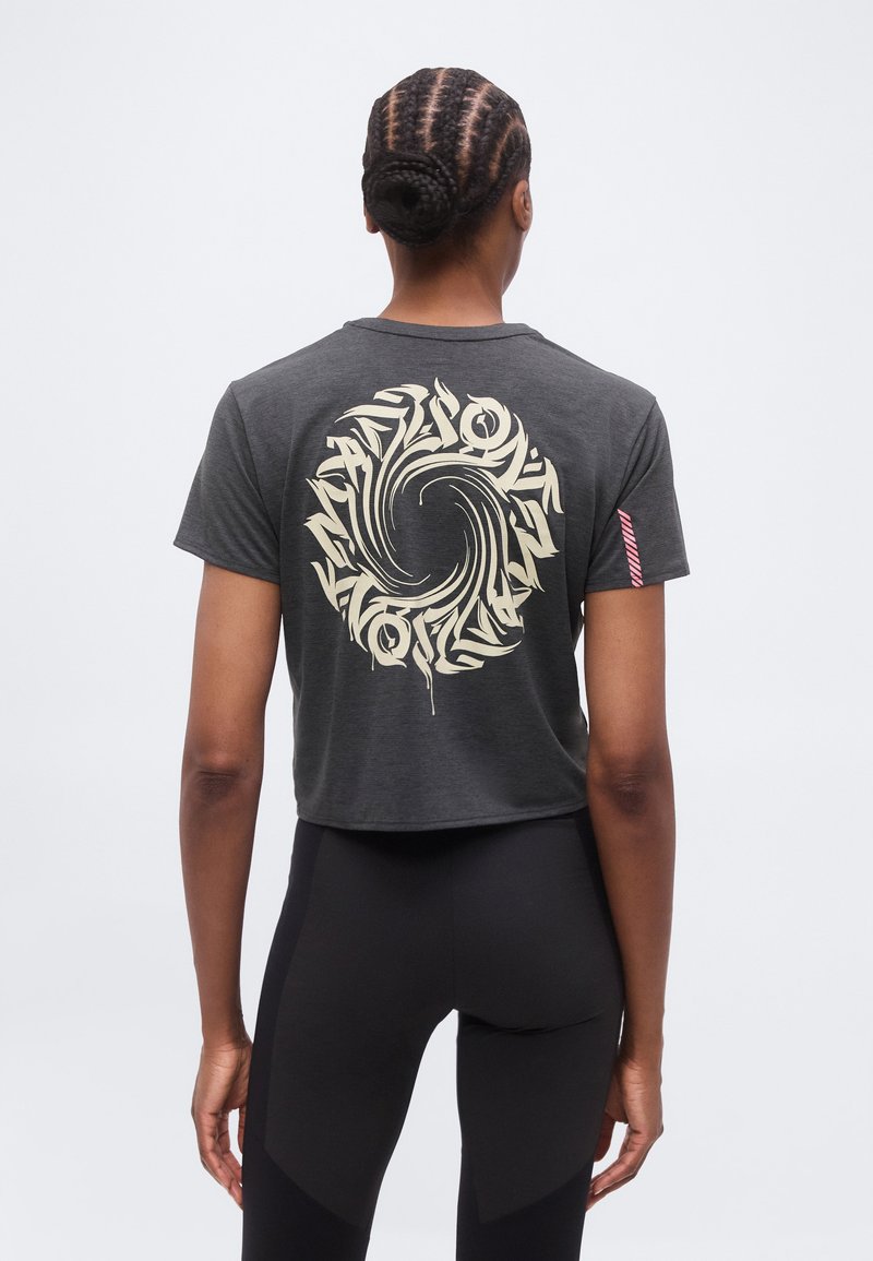 Γυναίκα που φαίνεται από πίσω, φοράει ένα σκούρο γκρι κοντό μπλουζάκι με κυκλικό, στροβιλισμένο κρεμ γραφικό στην πλάτη και μαύρο κολάν.