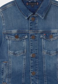 Giacca in denim blu con taglio classico, caratterizzata da due tasche sul petto, bottoni marroni e cuciture a contrasto. Colletto semplice e tessuto strutturato.