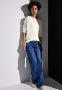 Stricktop in hellbeige mit kurzen, umgeschlagenen Ärmeln; kombiniert mit lockeren, blauen Jeans im geraden Schnitt und weißen Sneakern. Modernes, lässiges Outfit.