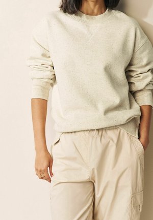 Personne portant un sweat crème à pois avec des poignets côtelés et un pantalon cargo beige, debout avec une main dans la poche devant un fond uni.