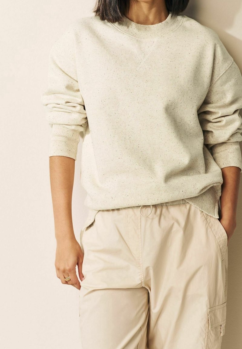 Personne portant un sweat crème à pois avec des poignets côtelés et un pantalon cargo beige, debout avec une main dans la poche devant un fond uni.