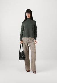Dark green knitted sweater with a high neck, paired with light beige wide-leg trousers. Model holds a black leather bag. Simple background.